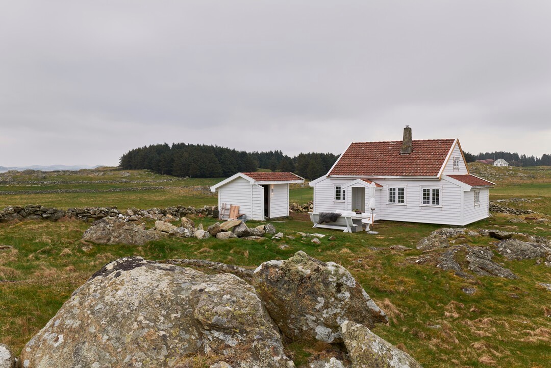 Eventyrlig oppussing, En hvitmalt hytte med klassisk liggende ULTIMALT-kledning og rødt tak, beliggende i et værutsatt landskap med grønne enger og steingjerder.