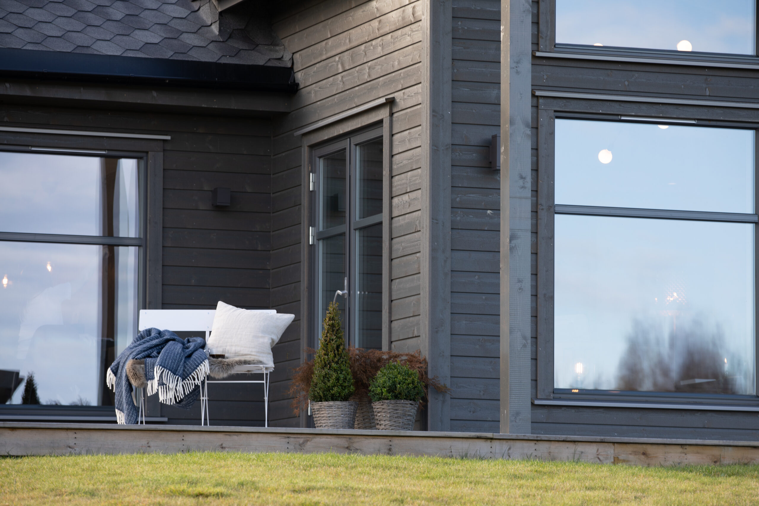 Fasade av moderne hus med liggende dobbelfals 60°-kledning i grå farge, store vinduer og utebenk med pledd og pynteputer.